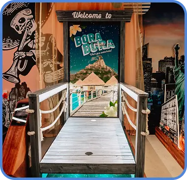 A wooden boardwalk putting green leading toward a vibrant "Welcome to Bora Bora" backdrop featuring overwater bungalows at a Hey Caddy indoor mini golf venue.