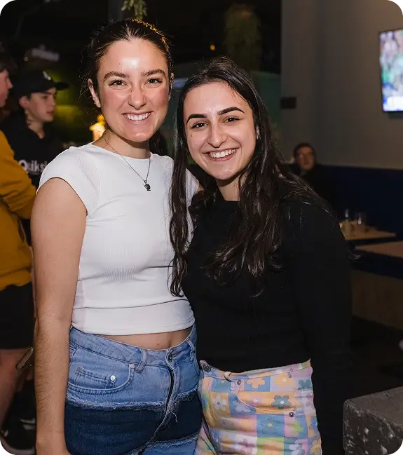 Two young women smiling and posing together during a casual night out at a lively indoor social venue and bar.