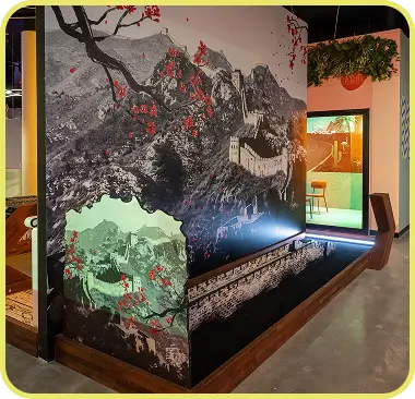 An indoor mini golf hole at Hey Caddy themed around the Great Wall of China, featuring a large mountain mural with red blossoms and a miniature replica of the wall as a putting obstacle.