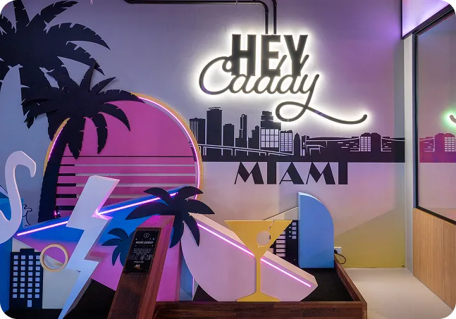 Miami-themed indoor mini golf hole at Hey Caddy featuring neon décor, tropical elements, and the iconic Hey Caddy signage.
