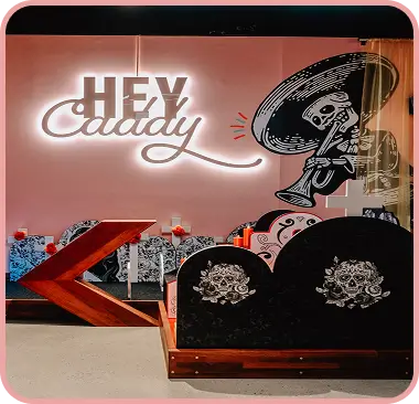 A brightly lit white neon Hey Caddy sign on a pink wall next to a Day of the Dead skeleton mural and sugar-skull-themed mini golf obstacles.