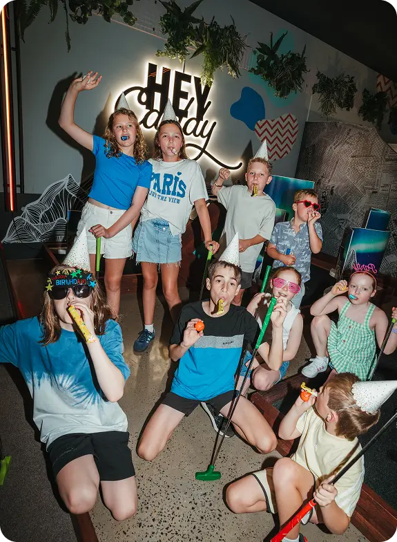 A group of excited children wearing party hats and sunglasses, blowing party horns, and holding colorful golf putters while celebrating a birthday on an indoor mini golf course under a glowing Hey Caddy neon sign.