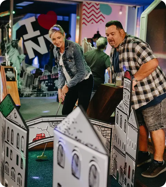 A stylized graphic banner for Hey Caddy mini golf featuring a woman walking down a long putting path toward a global city skyline and a Ferris wheel, alongside a man holding a cocktail against a mountain backdrop.
