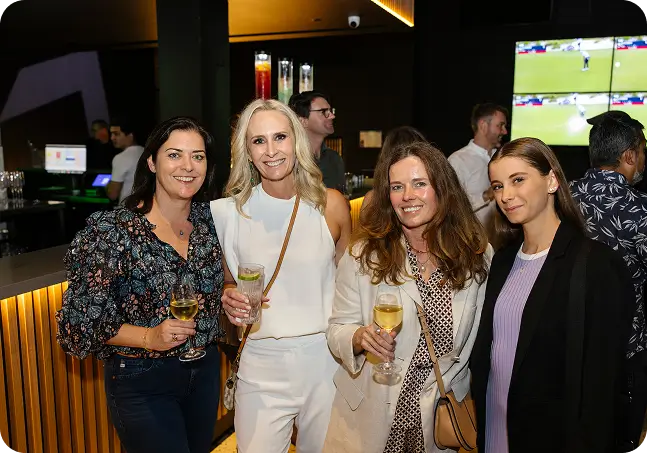 Four women smiling and holding drinks like wine and cocktails during a night out at a modern bar venue with golf simulator screens visible in the background.
