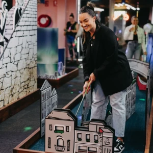 A smiling woman lining up a shot past mini city building obstacles on an indoor mini golf course at Hey Caddy.