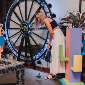 A woman lining up a putt on a mini golf course in front of a giant illuminated Ferris wheel backdrop at Hey Caddy.
