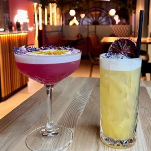 Two colorful handcrafted cocktails—a magenta coupe drink and a tall yellow highball garnished with edible flowers and dried citrus—sitting on a wooden table at Hey Caddy.