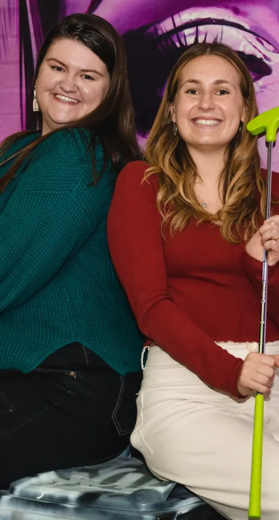 Two women smiling back-to-back while celebrating a Hens Party with a lime green golf putter at Hey Caddy.