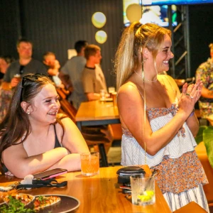 Two women smiling and clapping at a wooden table with cocktails and food platters inside the Hey Caddy lounge bar.