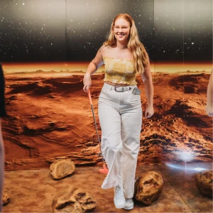 A smiling woman holding a putter walking across a Mars-themed indoor mini golf course with red rocky terrain murals and a starry sky backdrop at Hey Caddy.