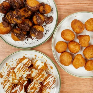 Three plates of golden mini donut bites topped with chocolate sauce, white chocolate drizzle, and classic glazes on a wooden table at Hey Caddy.