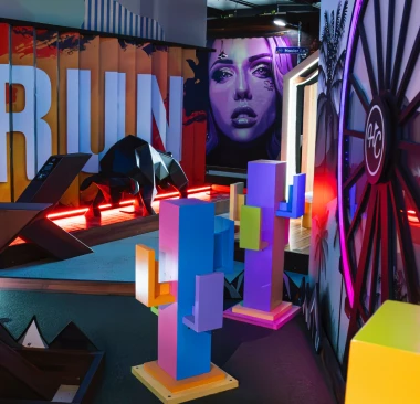 Hey Caddy mini golf hole featuring colorful geometric cactus obstacles, neon lighting, and a large wooden wagon wheel
