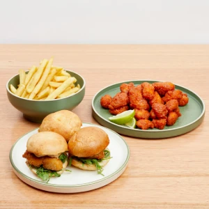 A food platter featuring three gourmet mini sliders, a bowl of crispy golden fries, and seasoned chicken bites with a lime wedge on a wooden table at Hey Caddy.