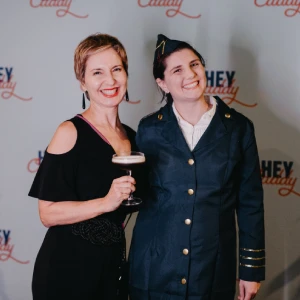 Two smiling women—one holding a cocktail and the other dressed in a pilot uniform—posing together in front of a Hey Caddy branded media wall backdrop.