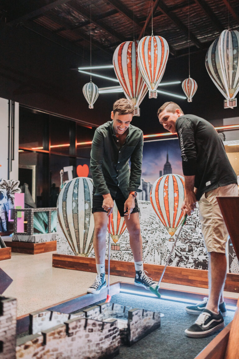 Two players enjoying indoor mini golf under balloon decorations at Hey Caddy Townsville
